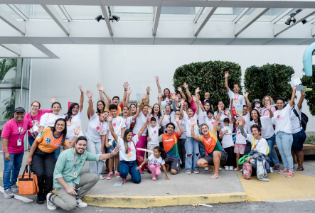 Grupo grande de pessoas sorrindo e acenando em um evento comunitário, com camisetas brancas e coloridas, em ambiente externo.